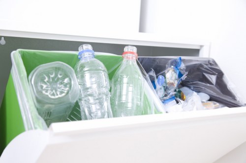 The image shows a white open storage drawer or bin filled with various disposable items, including two clear plastic bottles with blue and red caps, tilted on their sides and partially visible. Behind the bottles, there is a green plastic container or recycling bin that holds a clear, round plastic container with a lid, along with some other recyclable plastic waste. To the right, there is a black plastic bag filled with additional rubbish, possibly packaging materials or waste items, resting against the interior side of the drawer and slightly to the back. The environment appears to be a clean, indoor space, likely part of a food or waste disposal area, with bright, natural lighting highlighting the different textures and colors of the plastic items. This scene exemplifies typical rubbish items that Kentish Town House Clearance might manage in their waste removal services, illustrating the variety of plastic disposables and packaging waste involved in household or commercial clearances in the Kentish Town area.