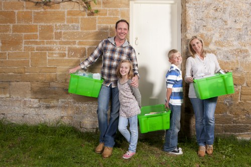A family of four stands outdoors against a weathered stone and brick wall, each holding a bright green plastic storage bin related to rubbish or waste disposal. The father, dressed in a casual checked shirt and jeans, is smiling while holding a bin in his right hand. A young girl with long hair and a grey sweater is standing next to him, also smiling and holding a bin. To their right, a boy in a striped shirt is partially visible, holding a bin and looking towards the camera. On the far right, a woman in a white blouse and jeans is holding a green bin, standing slightly angled with a neutral expression. The scene takes place on a grassy area adjacent to a paved space, with the family appearing to be engaged in rubbish or waste clearance activity, fitting for a housekeeping or rubbish removal service context in Kentish Town. The overall image suggests a family participating in a waste disposal task, with clear focus on the green bins and outdoor environment, suitable for referencing rubbish removal services in the local area.
