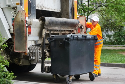 A waste collection worker dressed in high-visibility orange overalls and a white cap is standing outside on a paved area, operating a large waste compactor that is attached to a refuse collection vehicle. The worker appears to be using a control mechanism on the side of the compactor, which is positioned next to a dark grey wheeled waste bin with a textured surface, located on the right side of the image. The compactor, with its metallic rollers and mechanical components, is in the process of emptying or compacting waste from the bin. The background shows a green landscape with trees and a residential street, indicating a suburban or urban environment typical of the Kentish Town area. The scene is lit with natural daylight, emphasizing the industrial nature of rubbish collection services provided by Kentish Town House Clearance, located near the postcode area of NW5. This image illustrates the practical aspect of local refuse collection methods during routine rubbish removal operations.