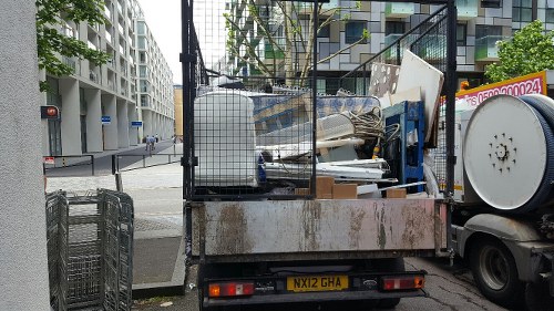 A flatbed waste collection truck parked on a city street with a black metal cage-like structure at the rear, filled with various types of rubbish including white and blue plastic debris, wooden panels, and metal objects, some stacked and leaning against each other. The truck is positioned near a pavement with a modern residential building with multiple balconies and large windows visible in the background, indicating an urban environment. The truck's surface appears weathered with visible rust and dirt, and the vehicle's license plate reads 'MX12 GHA'. The scene suggests a rubbish removal service, likely from Kentish Town, as part of a house or property clearance, handled by Kentish Town House Clearance. Overhead, electrical lines cross the scene, and the area appears clean despite the presence of waste material ready for disposal or recycling.